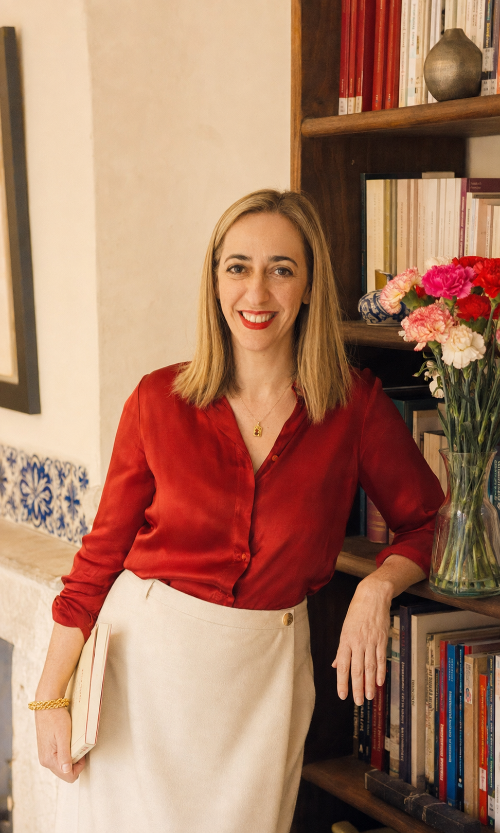 Carolina standing beside her bookshelves, holding a book.